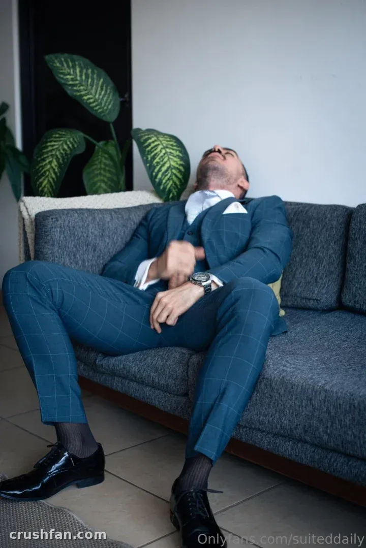 Man in a teal three-piece suit reclining on a grey sofa beside a large green plant