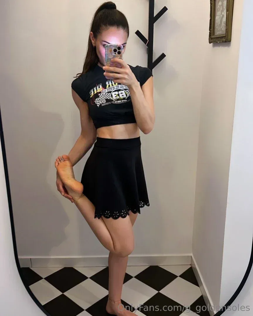 Mulher com um top cropped e saia pretos tirando uma selfie no espelho em uma sala com piso xadrez.