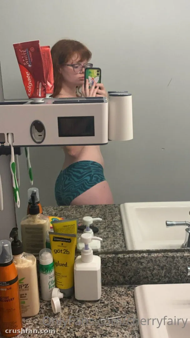 Cherry in glasses takes a mirror selfie in a bathroom with a cluttered counter.