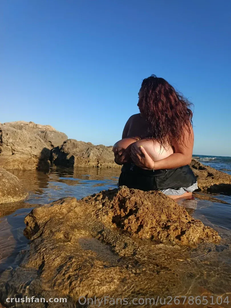 Hermosa сидит на камнях у воды под ясным голубым небом