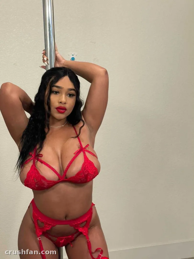 Thebae Breanna in red lace lingerie posing beside a metal pole against a light wall