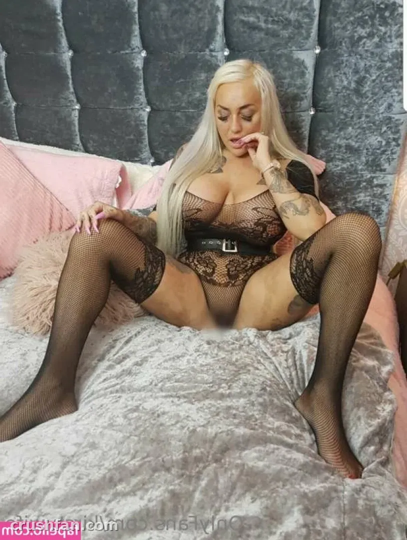 Creadora con body de encaje negro y medias de rejilla sentada en una cama gris con cojines rosas