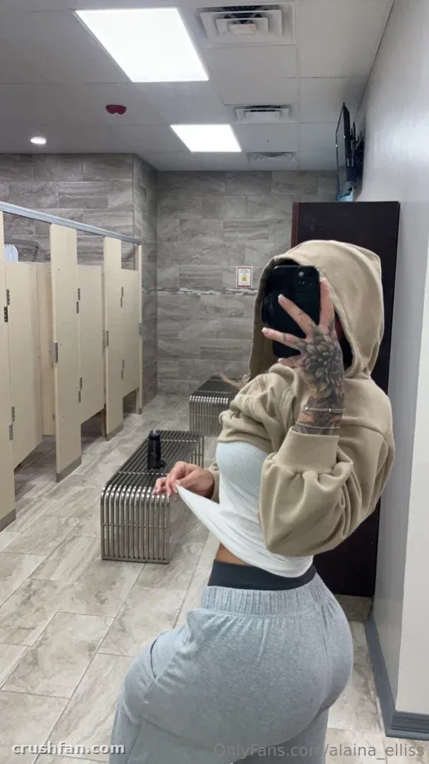 Alaina in a beige hoodie and grey sweatpants taking a mirror selfie in a tiled public restroom