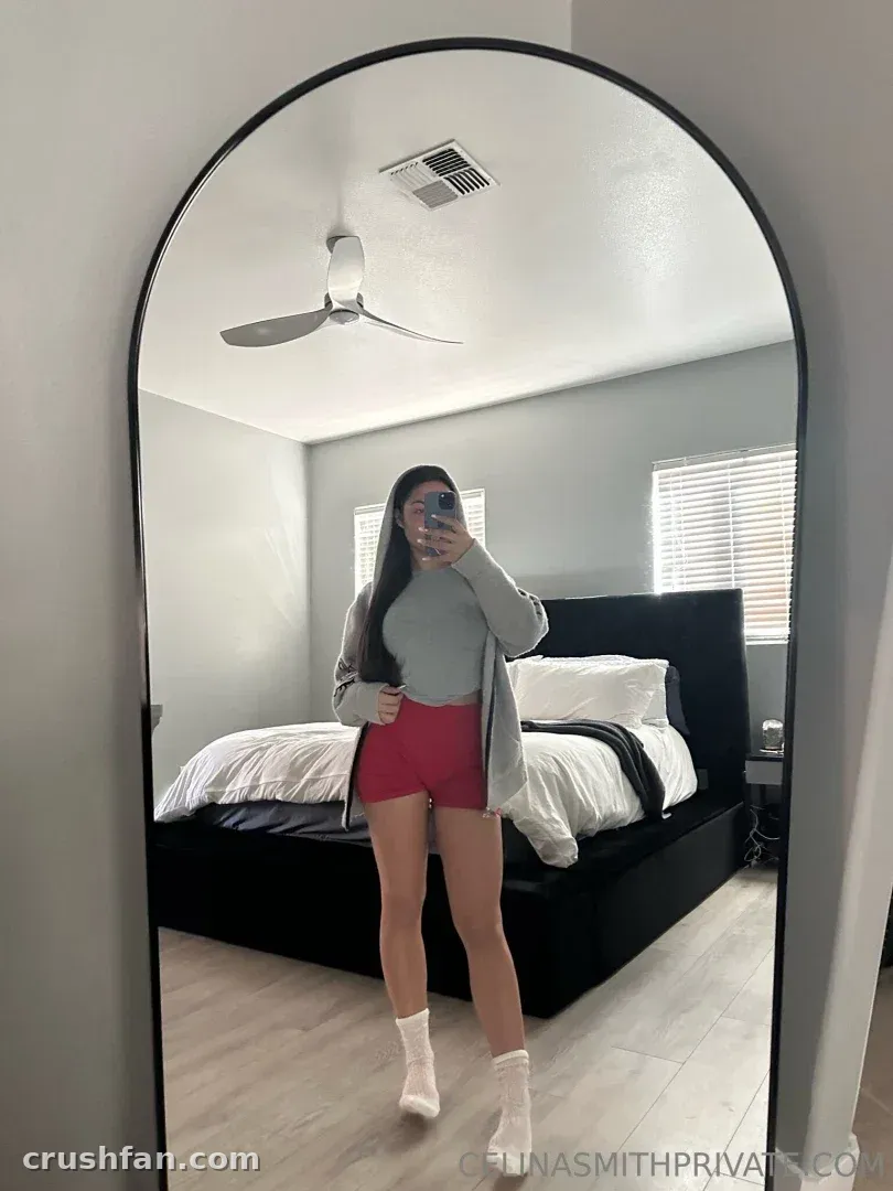 Cel con una parte superior gris y pantalones cortos rojos tomándose un selfie frente al espejo en un dormitorio luminoso.