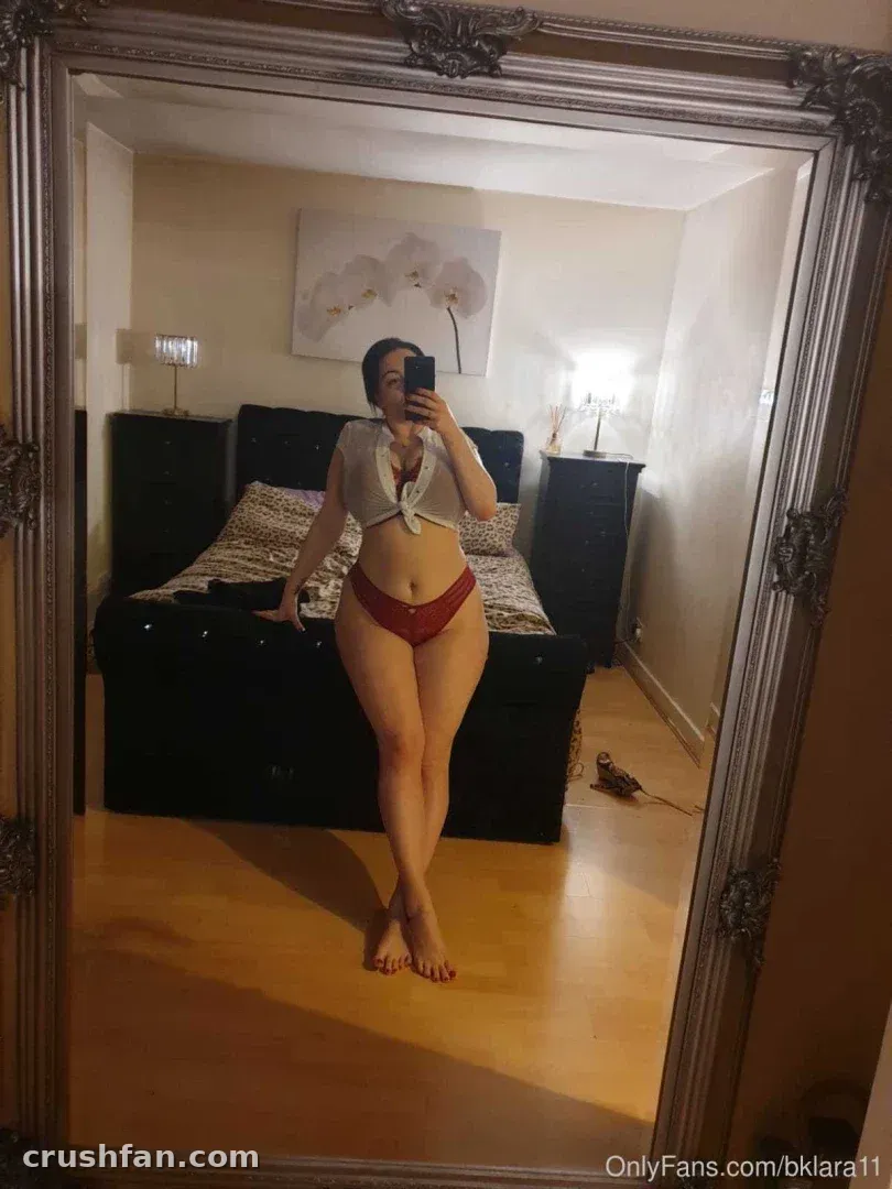 Woman in a light top and red bottoms taking a full-length mirror selfie in a warm bedroom.