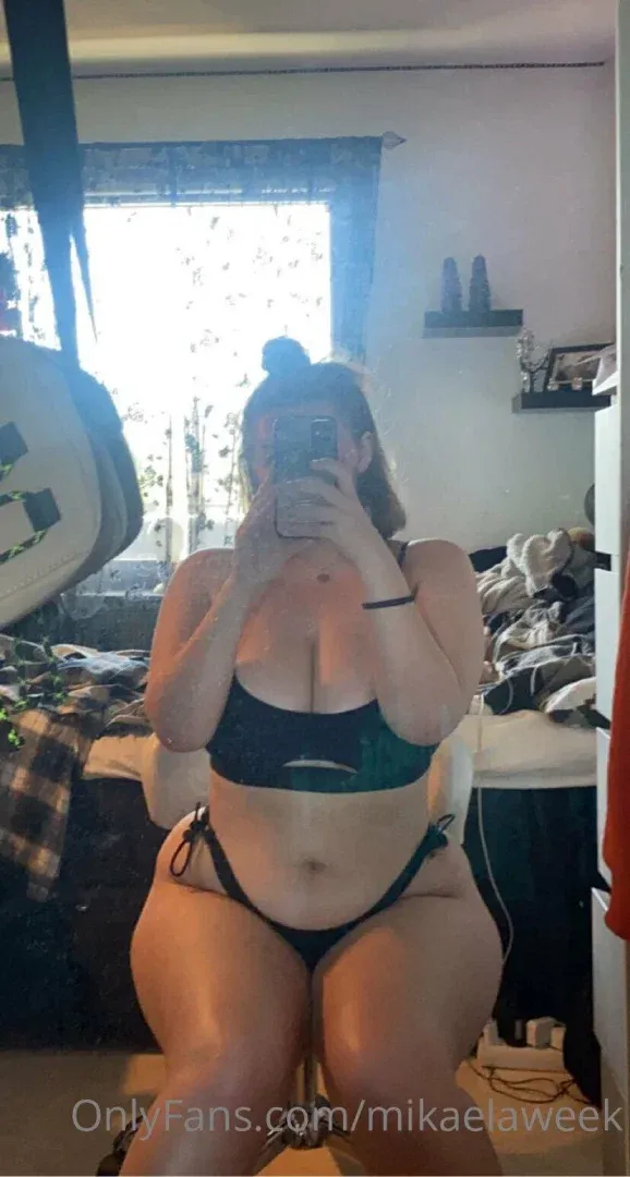 Mika22 prenant un selfie dans le miroir en bikini vert dans une chambre avec une fenêtre lumineuse derrière elle.