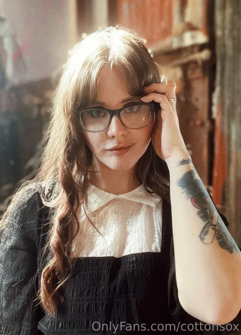 Alice CottonSox in glasses and a white collared blouse with a tattooed arm, warm indoor light.