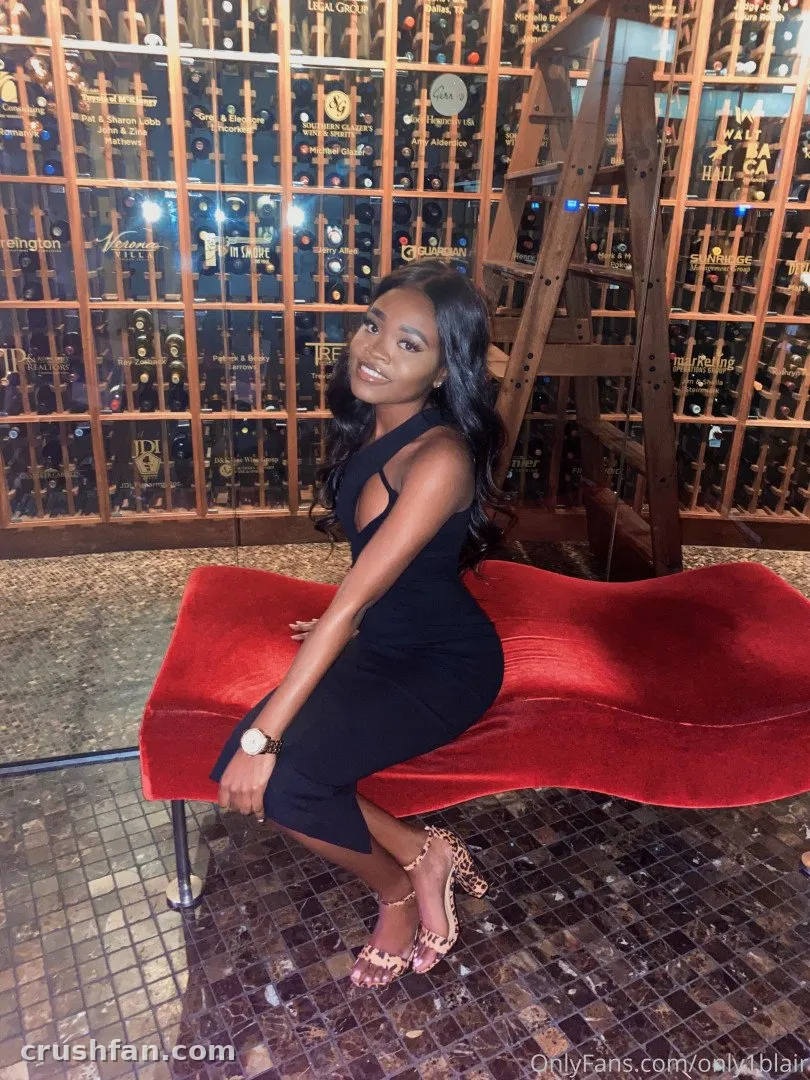 Blair in a black dress sitting on a red velvet bench in front of a wooden wine rack