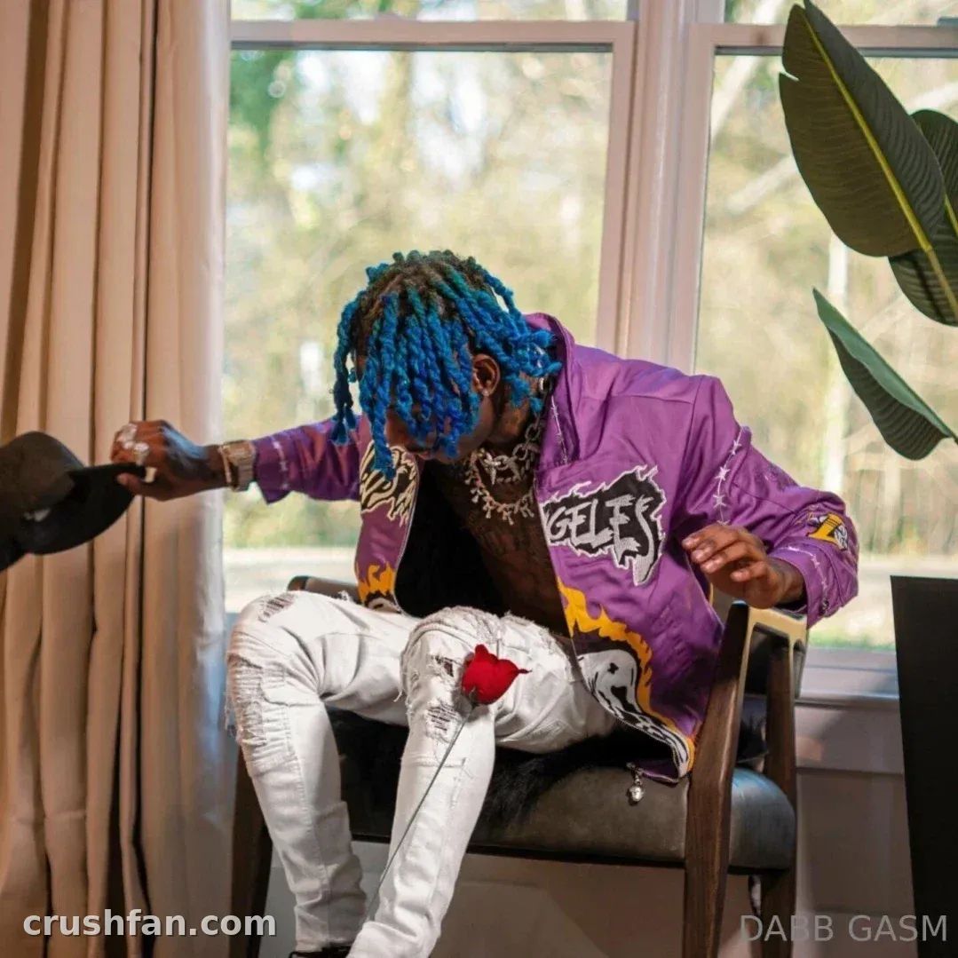 DABB LAND seated indoors in a purple jacket and white jeans holding a hat