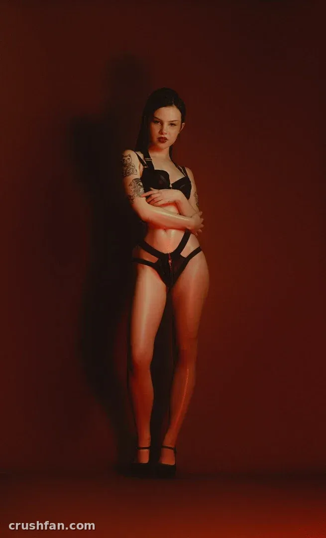 Gwen em lingerie preta e saltos altos de pé diante de uma parede vermelho intenso