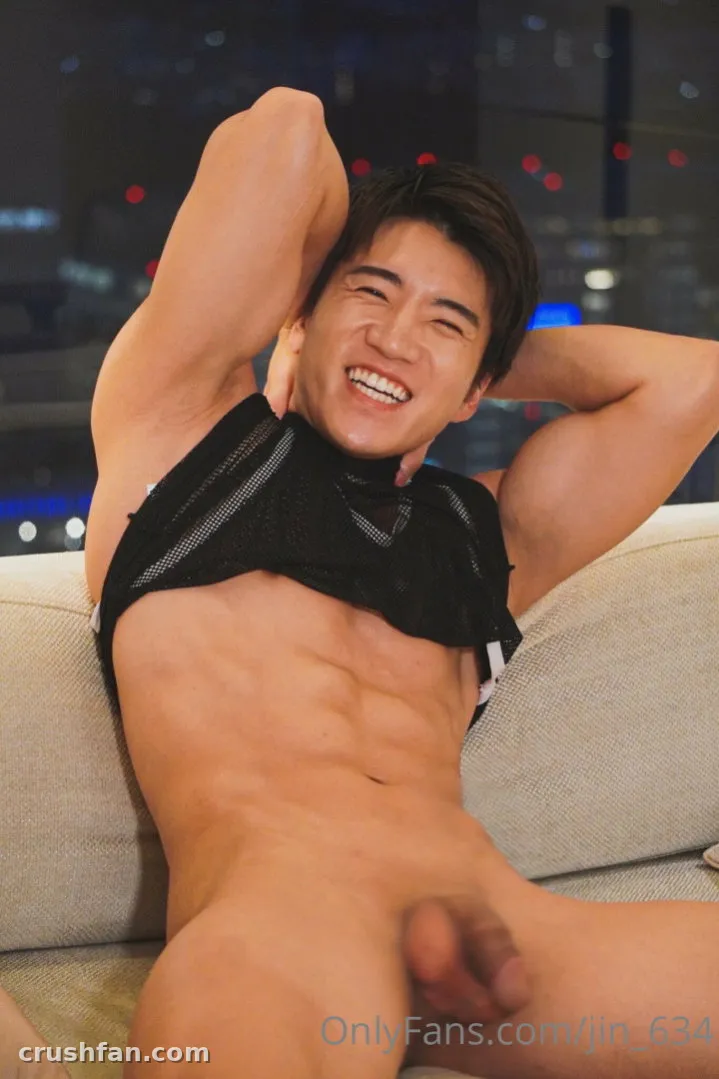 Smiling man in a black mesh top reclining on a beige sofa with city lights behind him