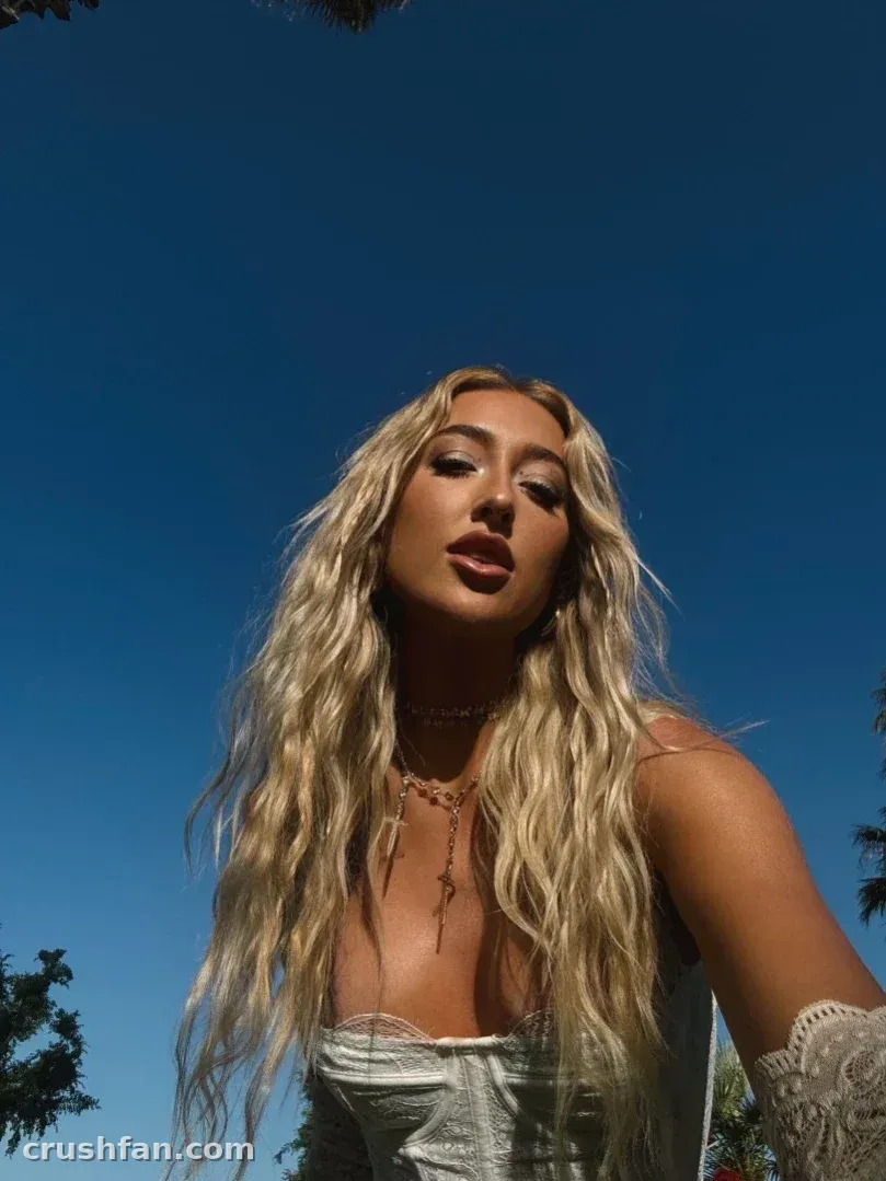 Madds com longo cabelo loiro ondulado ao ar livre contra um céu azul profundo, vestindo uma blusa de renda branca.