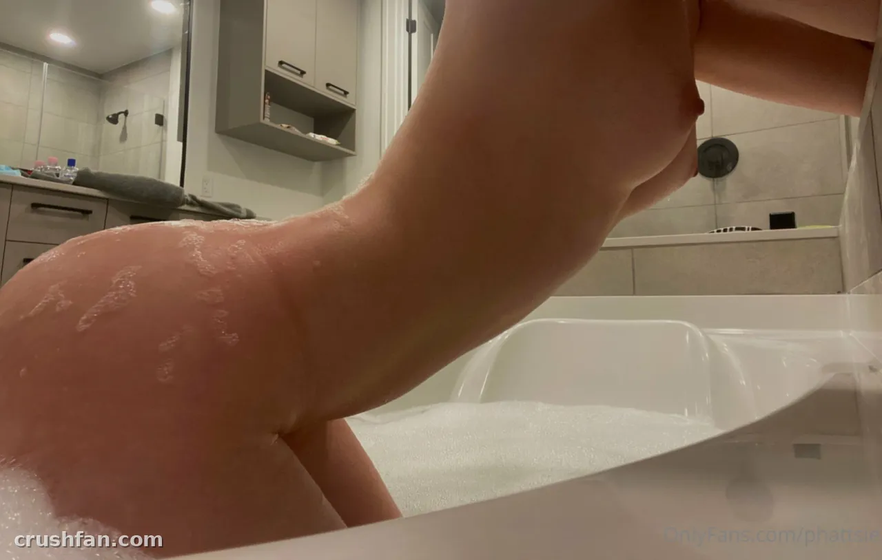 Emi leaning over a white bathtub with foam in a bright tiled bathroom