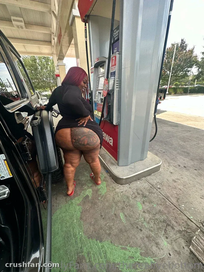 Fyebottom in einem schwarzen Outfit und roten High Heels, die an einer Zapfsäule einer Tankstelle posiert.