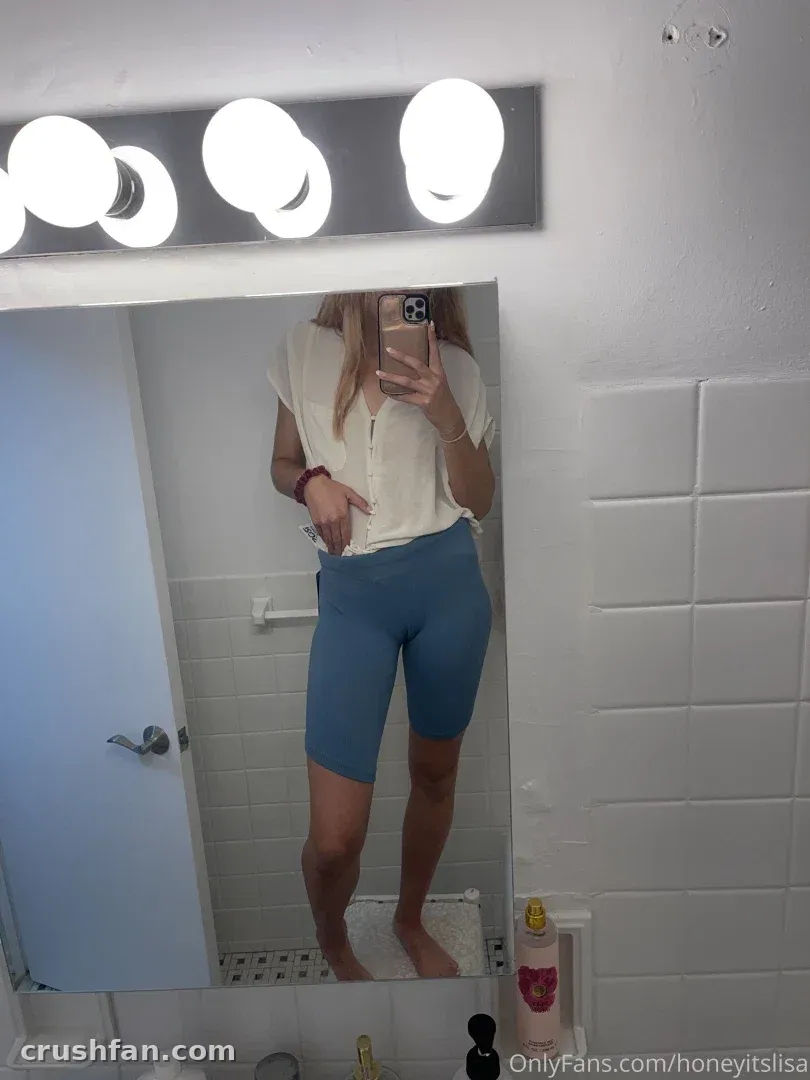 Lisa tirando uma selfie no espelho em um banheiro claro, usando uma blusa clara e short azul.
