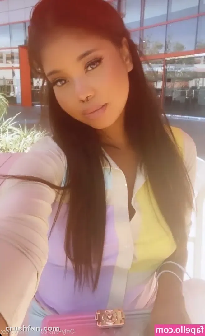 Nia Nacci com uma blusa de botões pastel e jeans tirando uma selfie ao ar livre à luz do dia