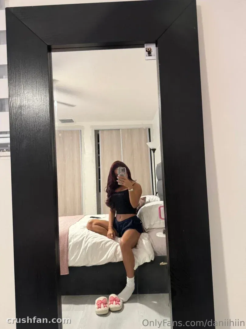 Dani taking a mirror selfie in a black top and shorts in a bright bedroom