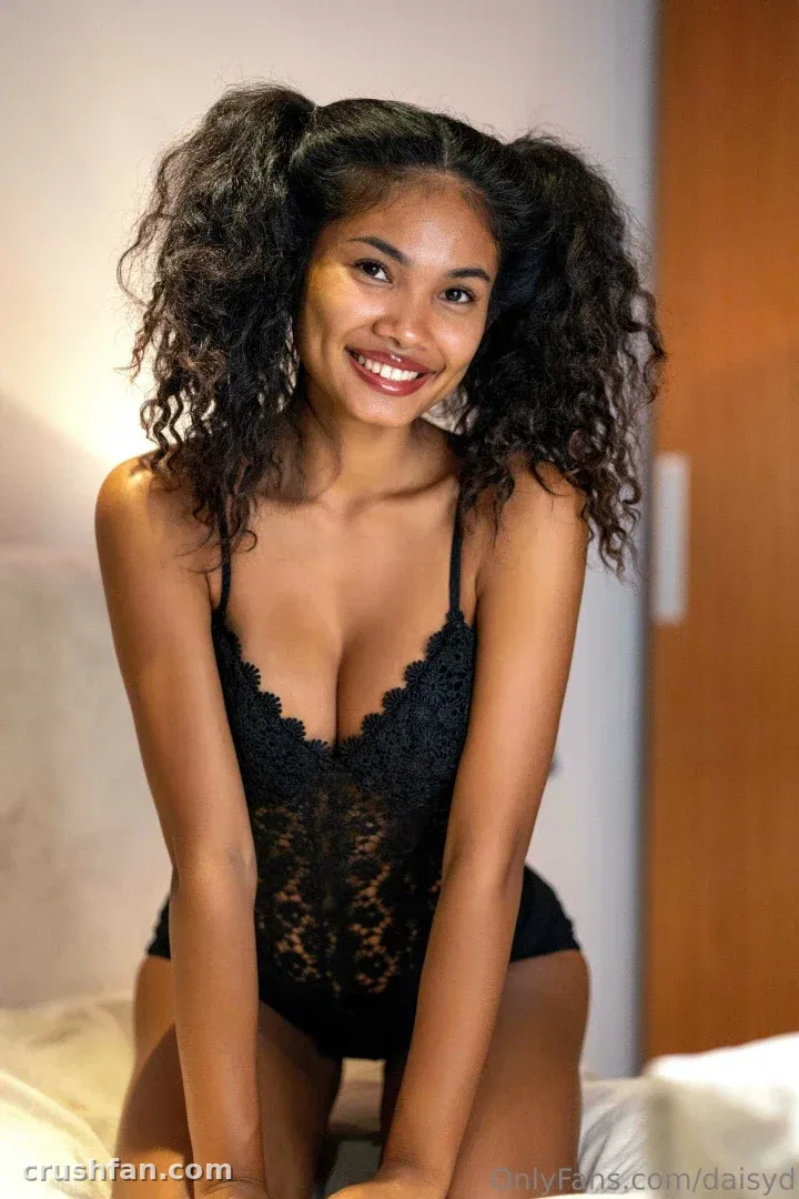 Daisyd sorrindo com um body de renda preta e marias-chiquinhas cacheadas em um quarto com luz suave
