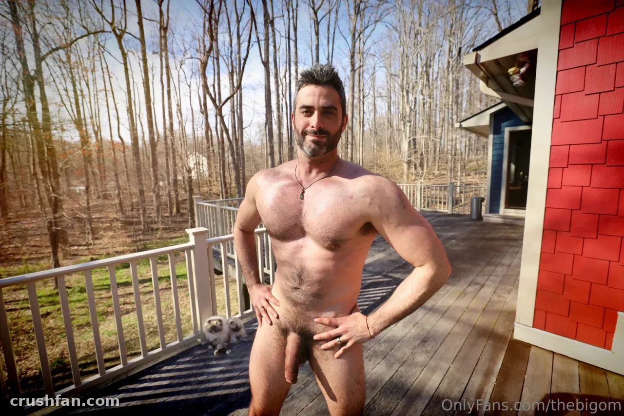 Nude man with a pendant necklace posing on a sunlit wooden deck beside a grey cat and red-sided building