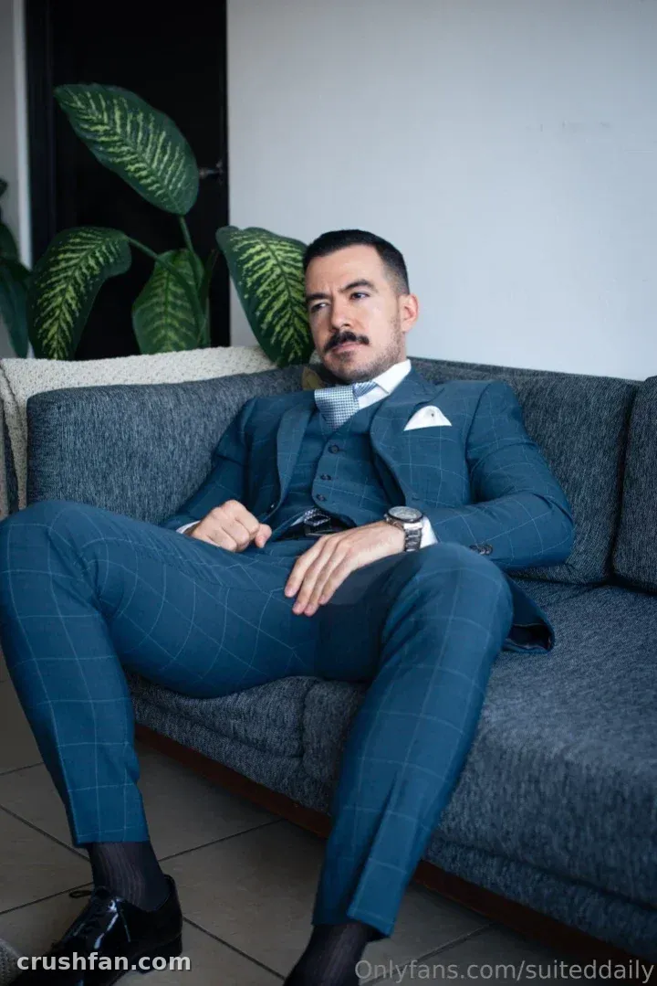 Creator in a teal checked three-piece suit seated on a grey sofa indoors