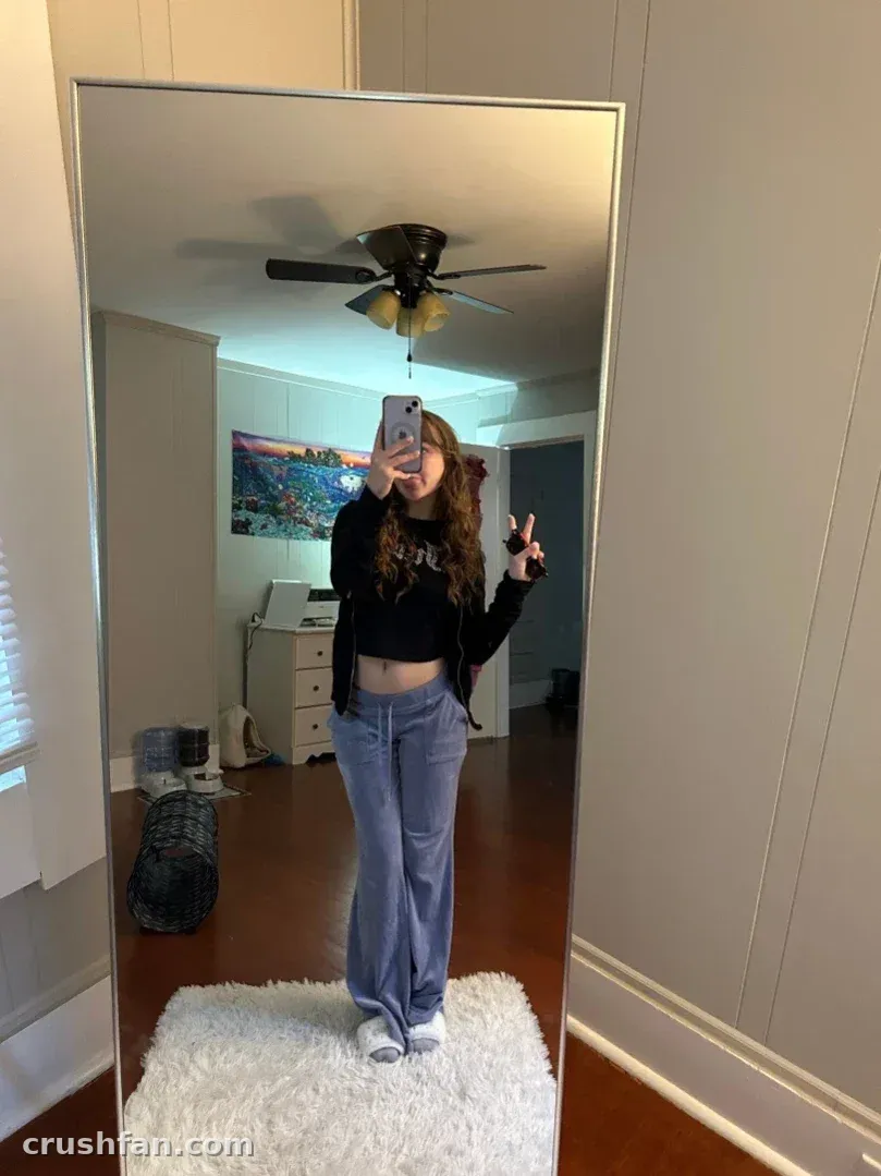 Kali in a black top and blue jeans taking a mirror selfie in a bright room with a cream rug