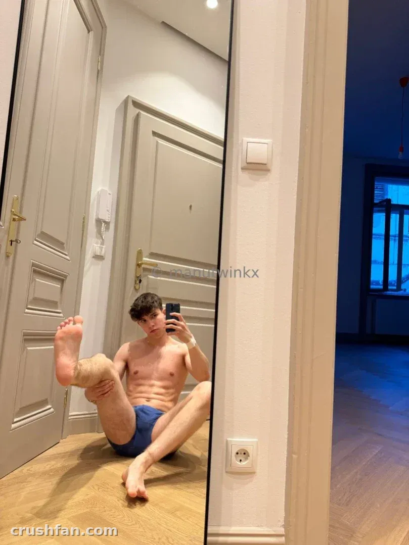 Manu está sentado em um piso de madeira tirando uma selfie no espelho em um cômodo claro com portas brancas.