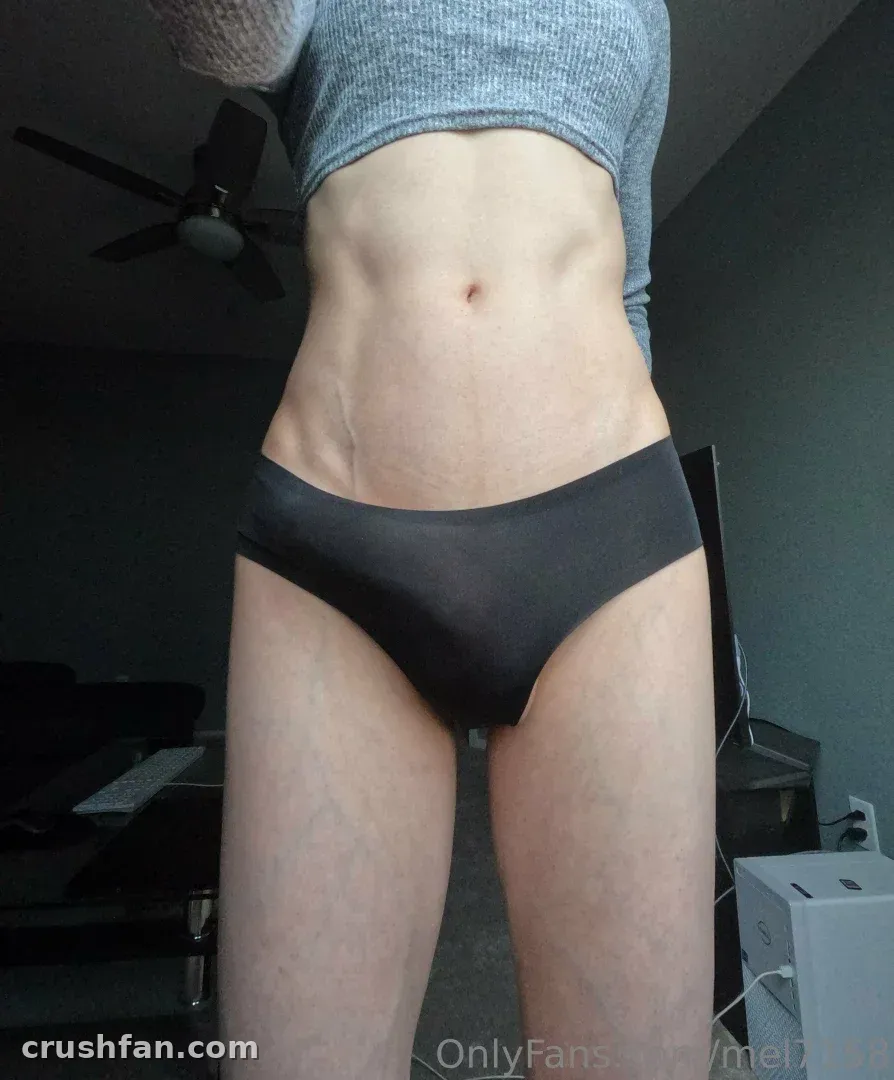 Melany en crop top gris et culotte noire debout dans une pièce intérieure faiblement éclairée