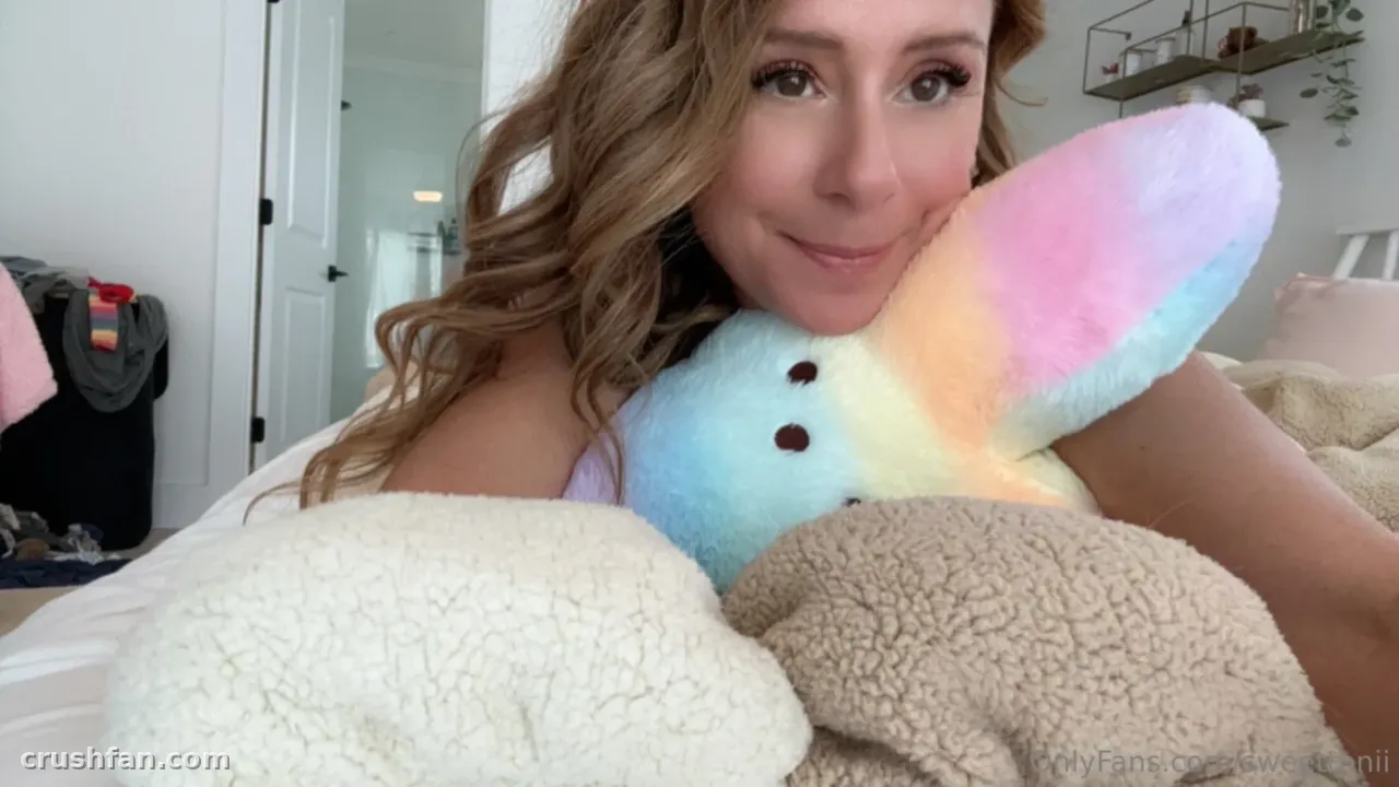 Sweettonii sosteniendo un peluche de arcoíris pastel en una cama con cojines suaves.