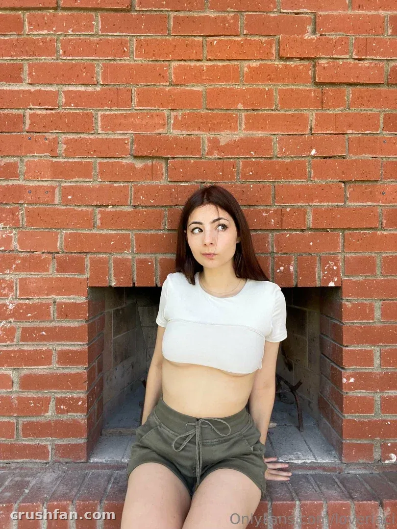 Laci Witton dans un crop top blanc et un short olive assise près d'un mur de briques rouges