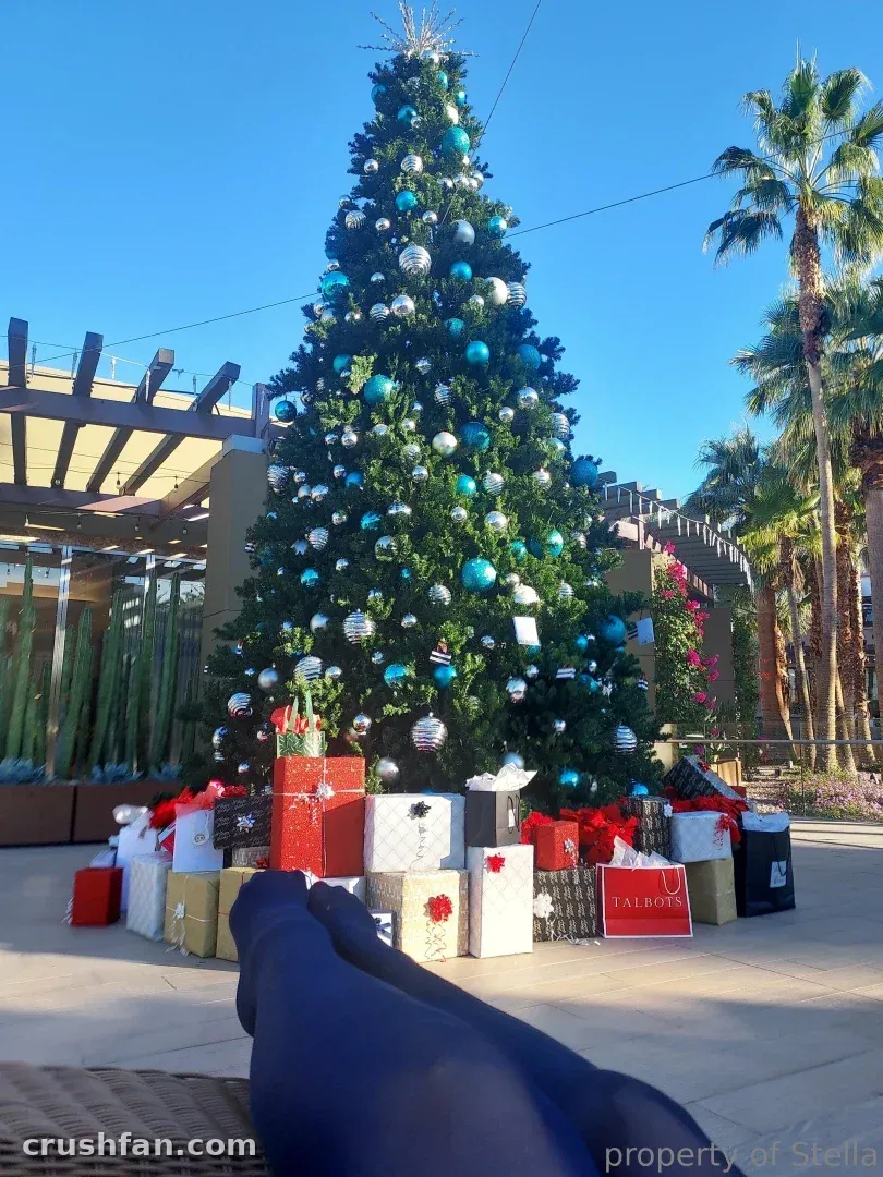 Pernas com meia-calça escura e opaca esticadas em frente a uma árvore de Natal decorada com presentes.