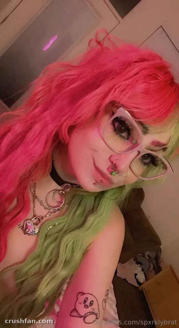 Selfie de perto de uma mulher com cabelo rosa e verde e óculos transparentes em ambiente interno