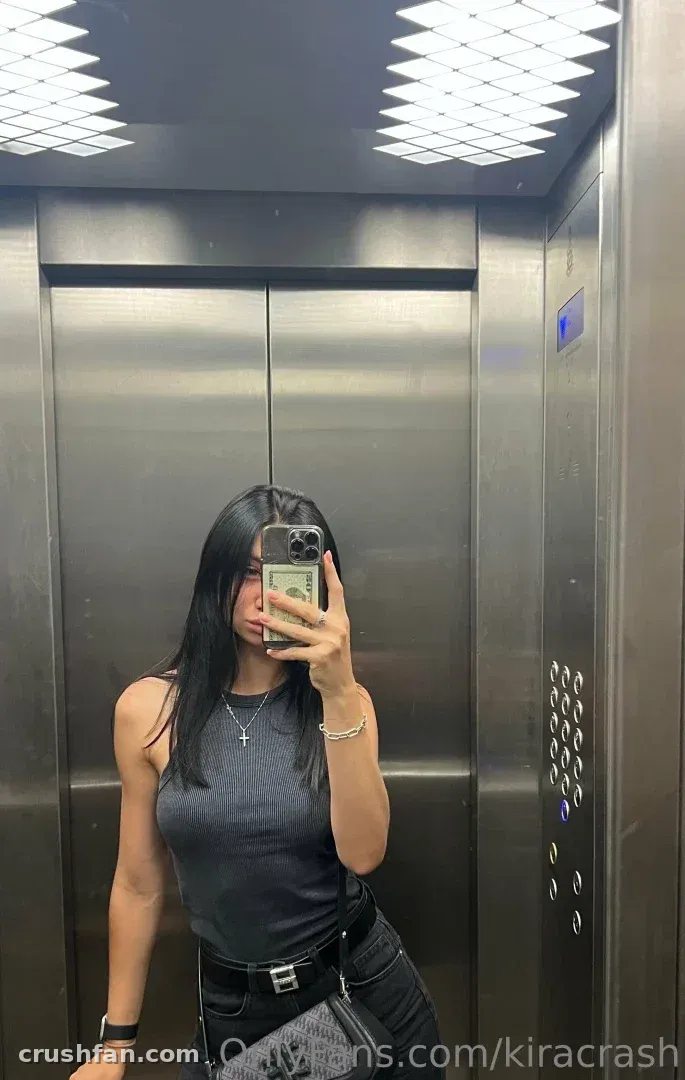 Kira con una camiseta sin mangas gris tomándose una selfie en el espejo dentro de un ascensor metálico