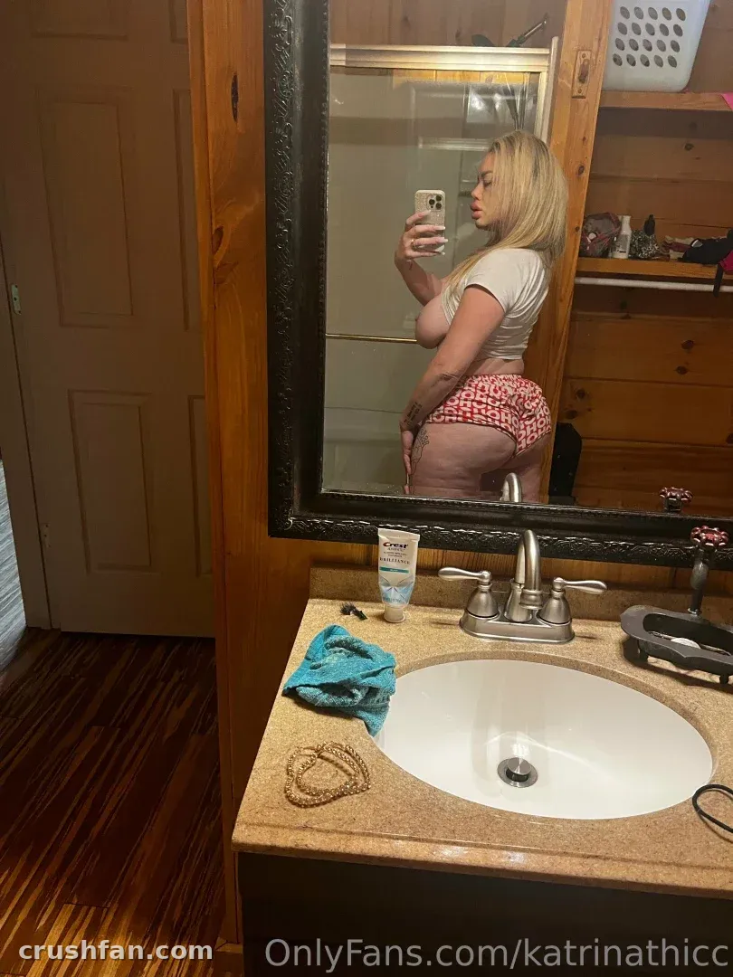 Katrina Thicc prenant un selfie dans le miroir dans une salle de bain, portant un haut blanc et un bas à motifs rouges.