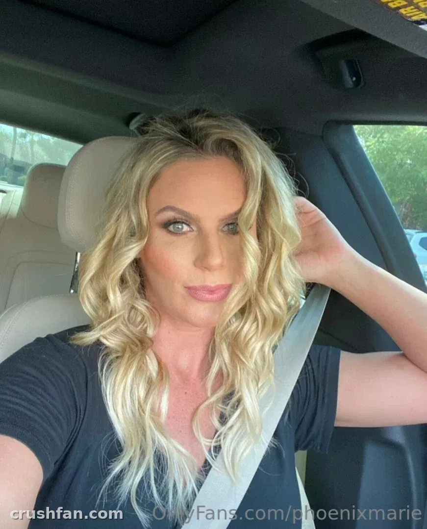 Mulher loira de blusa preta sentada em um carro, tirando uma selfie à luz do dia