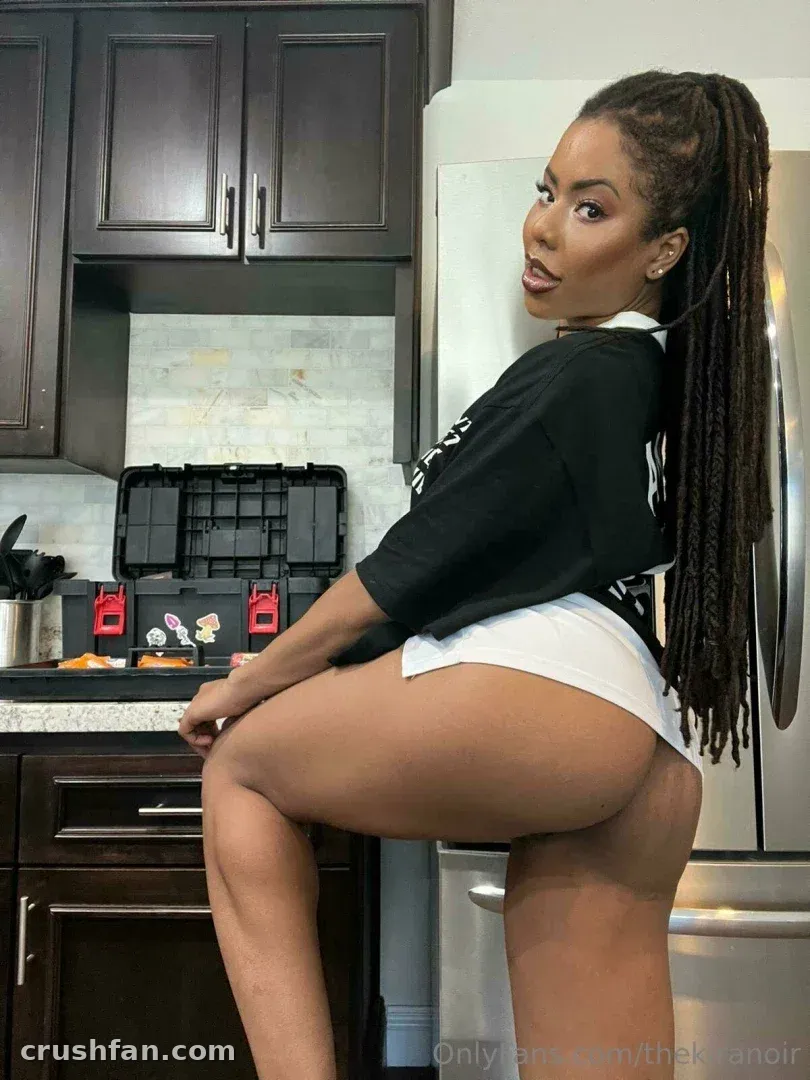 Kira Noir dans une cuisine portant un haut noir et un short blanc, regardant vers l'appareil photo.
