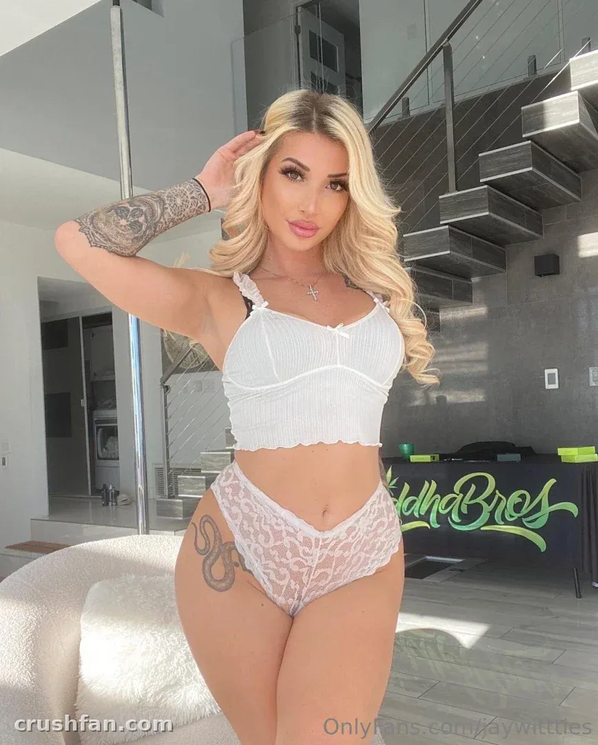 Mulher loira com top cropped branco e calcinha de renda posando em ambiente interno perto de uma escada