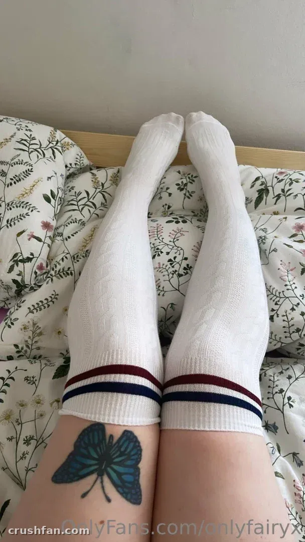 Pernas com meias brancas listradas até o joelho descansando sobre lençóis florais, com uma tatuagem de borboleta azul visível.