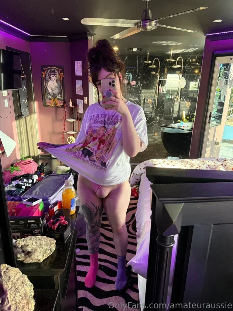 Aus Sarah tirando uma selfie no espelho em um quarto com luz roxa, usando uma camiseta oversized.
