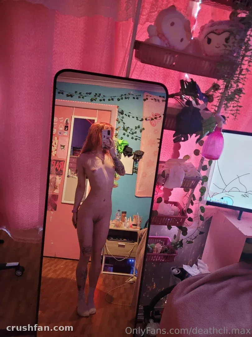 Maxine con un body claro tomándose una selfie de cuerpo entero frente al espejo en una habitación con luz rosa.