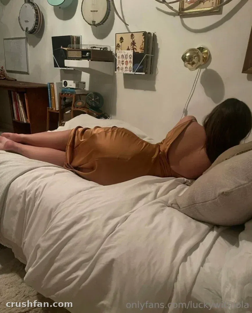 Luckywithlola lying on a bed in a bronze satin dress in a room with banjos on the wall
