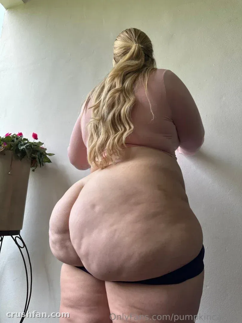 Pumpkin Cakezz está de pie en interiores con un top rosa y bragas oscuras junto a una planta en maceta