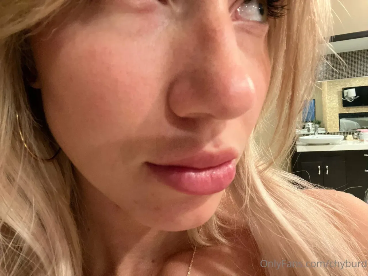 Close-up portrait of Chy Burd with blonde hair and hoop earrings in a bathroom.