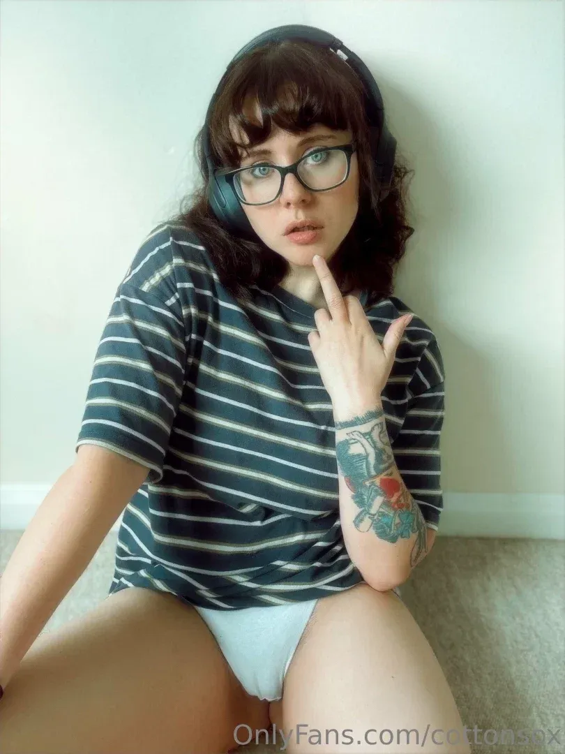 Alice CottonSox in a striped shirt, glasses and headphones sitting on a carpet indoors