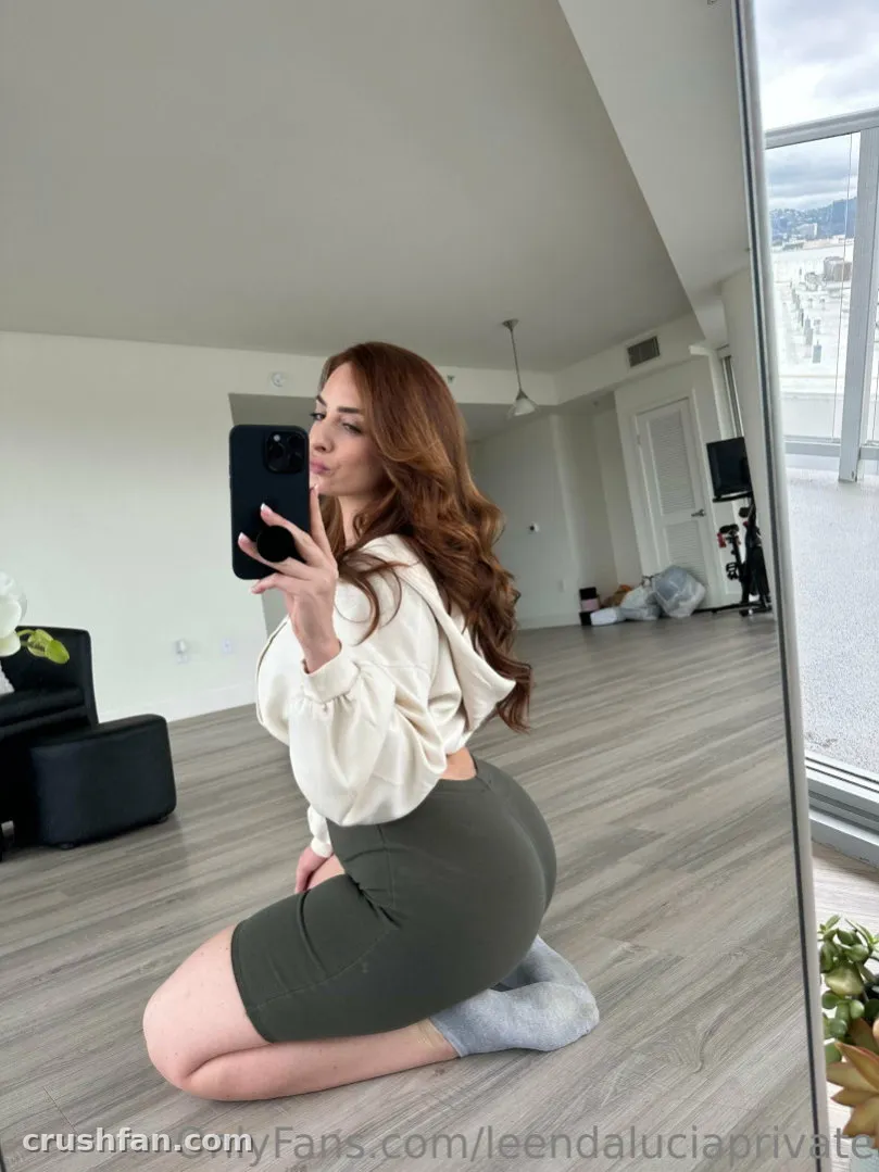 Leenda Lucia ajoelhada em um piso de madeira tirando uma selfie no espelho em um quarto claro