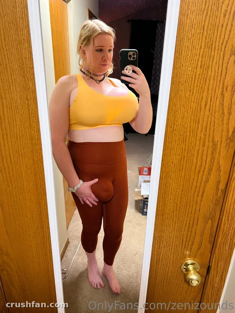 Criadora tirando uma selfie no espelho com top esportivo amarelo e leggings ferrugem junto a uma porta de madeira.