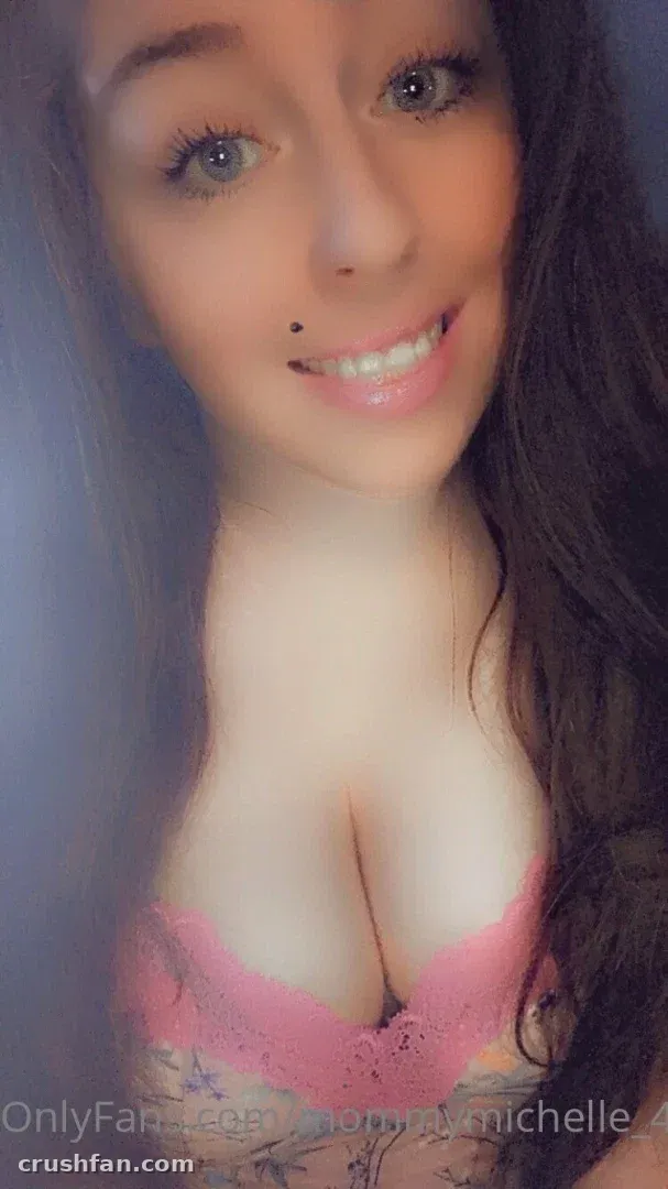 Michelle sonríe en una selfie cercana con una parte superior de encaje rosa sobre un fondo pálido.