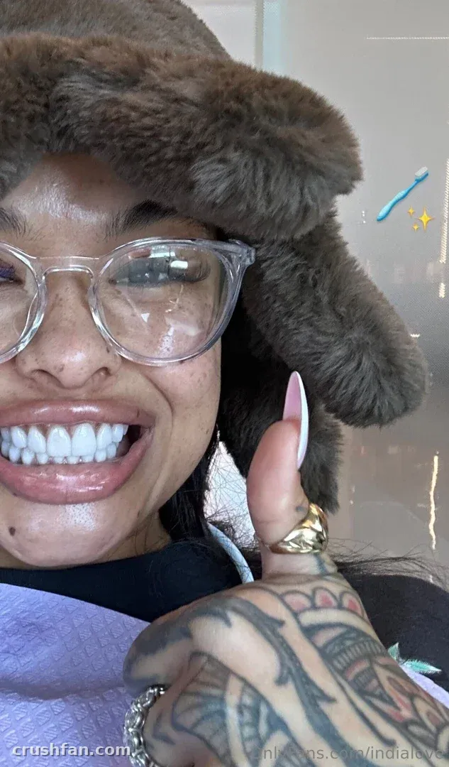 India Love sorri com um chapéu marrom fofo e óculos transparentes, levantando uma mão tatuada em direção à câmera.