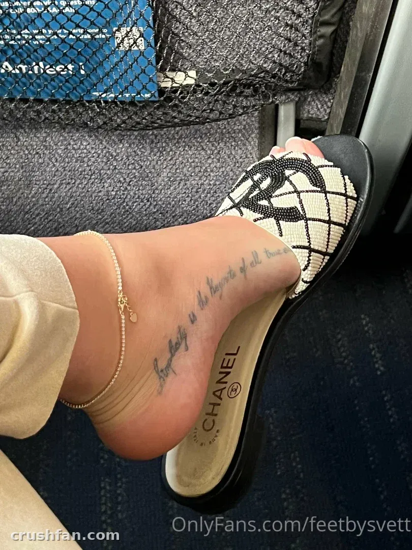 Close-up de um pé em um slide estampado em preto e branco com uma tornozeleira dourada e tatuagem em letra cursiva.