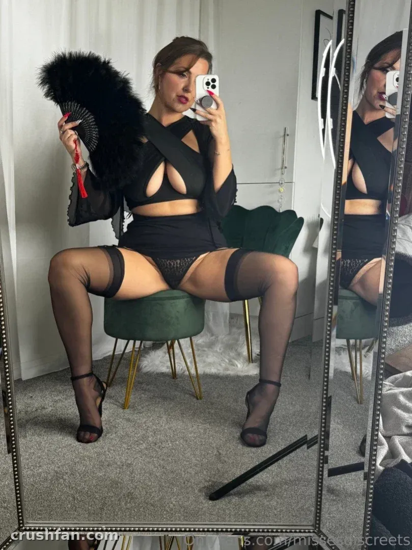 Woman in black outfit with feather fan taking a mirror selfie while seated on a green stool.