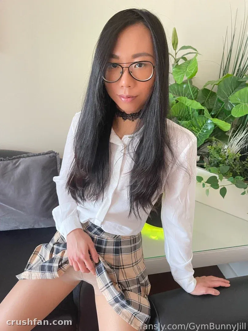 Jill in a white shirt and plaid skirt sitting on a sofa with a plant behind her